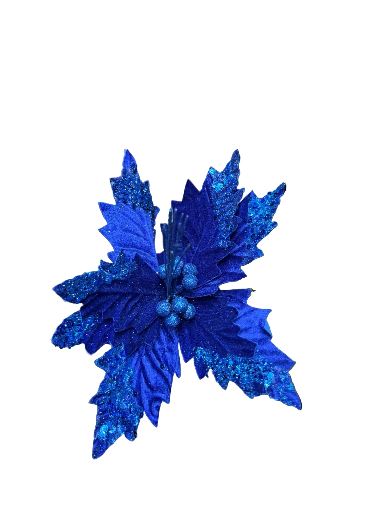 A close-up of a glittery royal blue artificial poinsettia flower with textured leaves and small berry-like accents, held in a hand. The background shows a cluttered workshop or studio environment with cardboard boxes, cables, and other decor items.