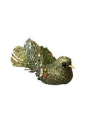 A hand holds a green glitter bird Christmas ornament with a metallic gold tail, in front of a red shopping basket filled with holiday decorations on a wooden floor.