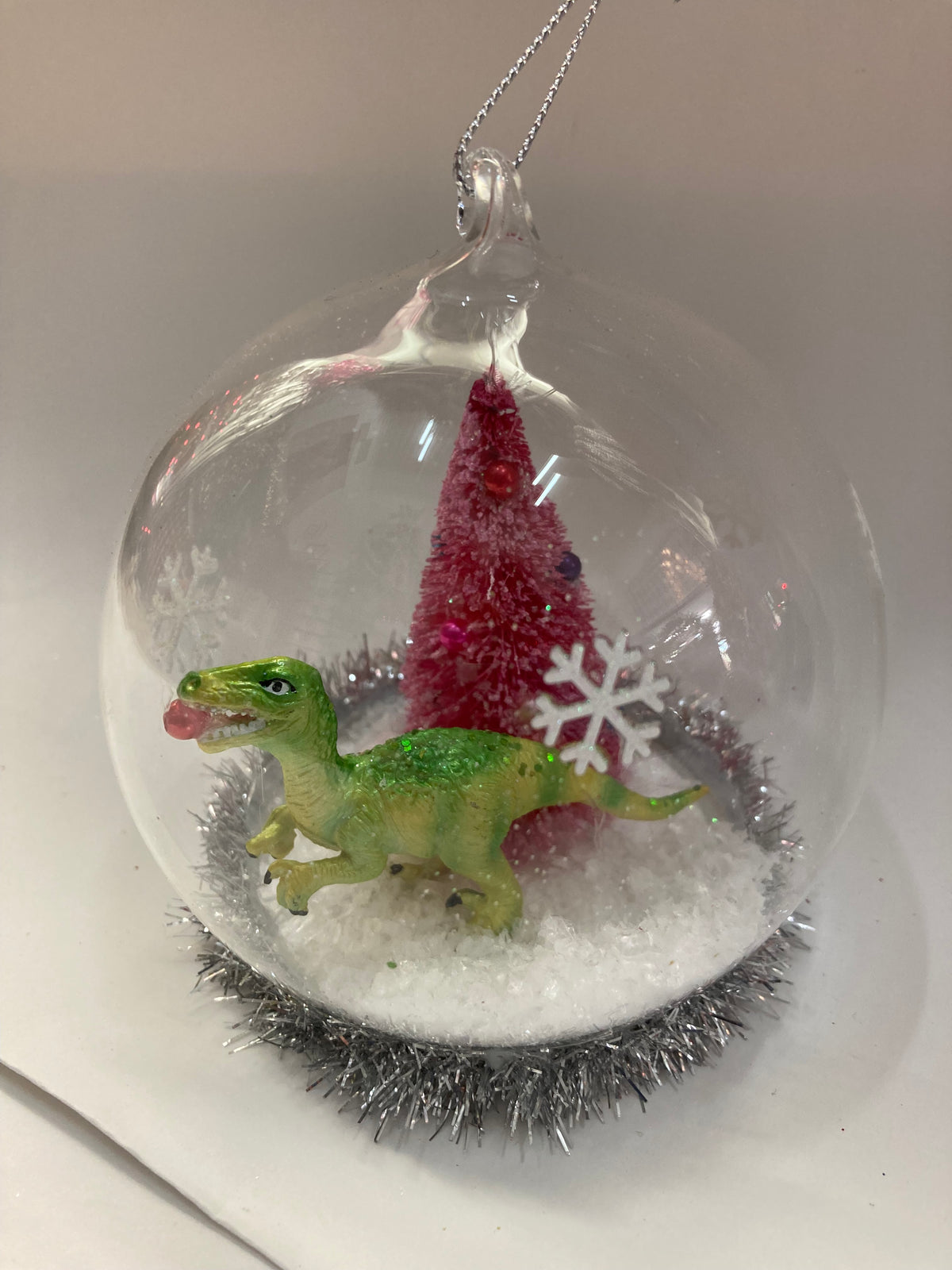 A clear glass Christmas ornament shaped like a snow globe, containing a small green dinosaur figurine, a red Christmas tree, a white snowflake, white faux snow, and a silver tinsel base, hanging by a clear loop.
