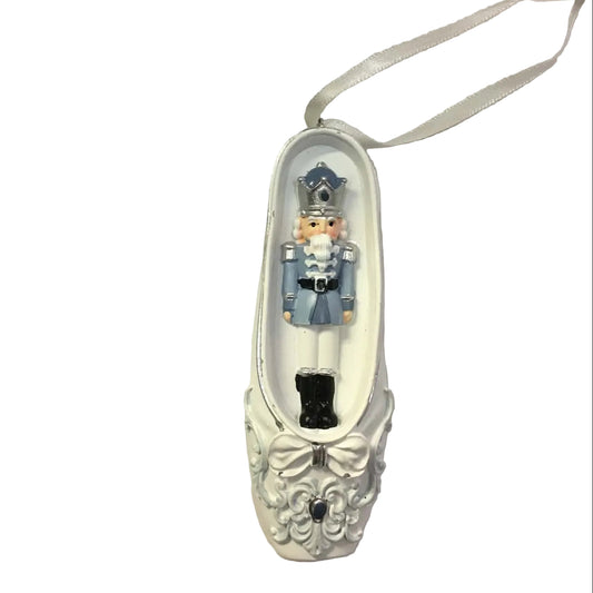 A pair of white ballet flat Christmas ornaments shaped like ballet shoes, with one containing a ballerina figurine and the other a nutcracker figurine. A black price tag is attached to the white ribbons.