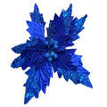 A close-up of a glittery royal blue artificial poinsettia flower with textured leaves and small berry-like accents, held in a hand. The background shows a cluttered workshop or studio environment with cardboard boxes, cables, and other decor items.